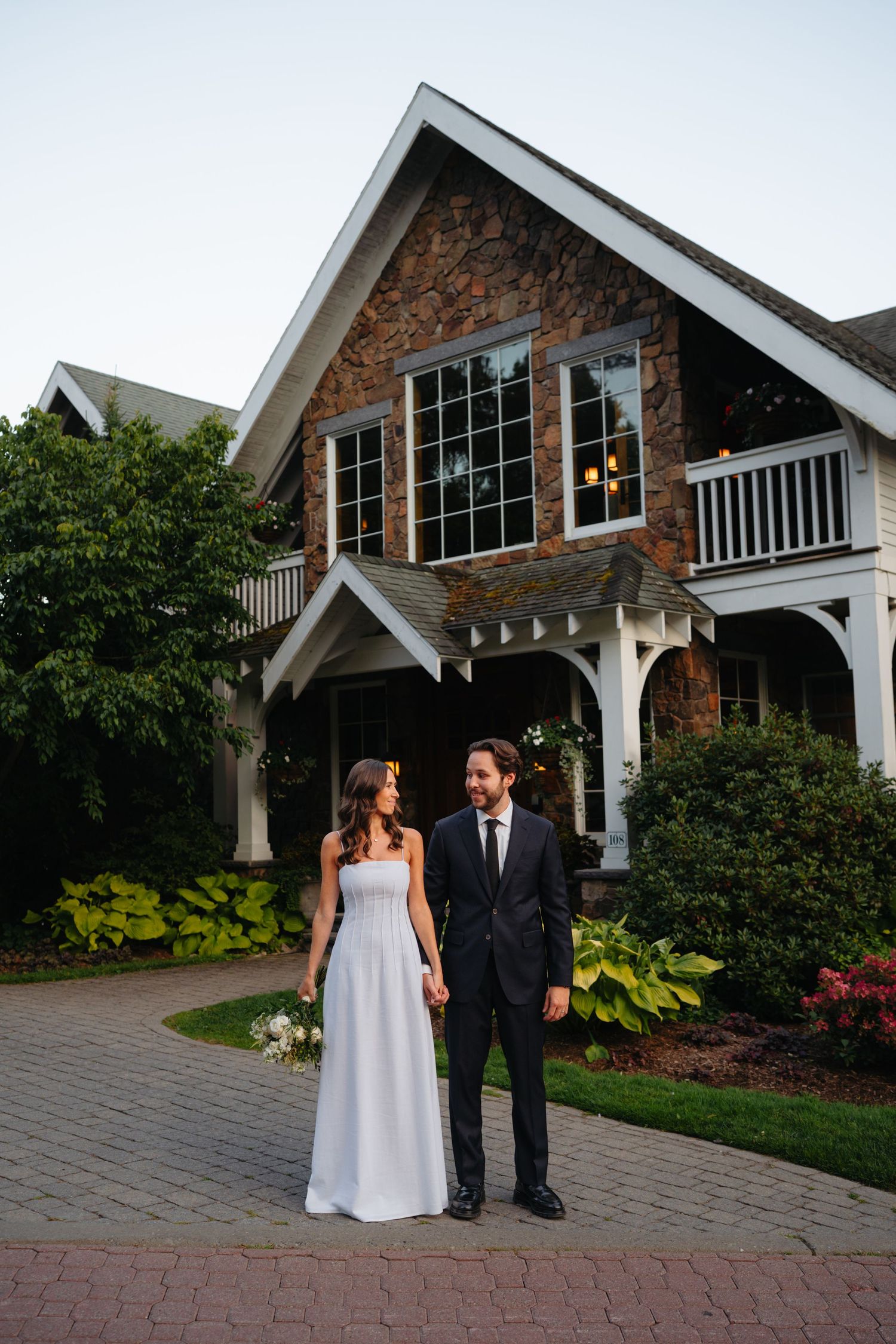 Bride and groom hold hands in front of a charming stone and white wood venue surrounded by lush greenery at dusk.