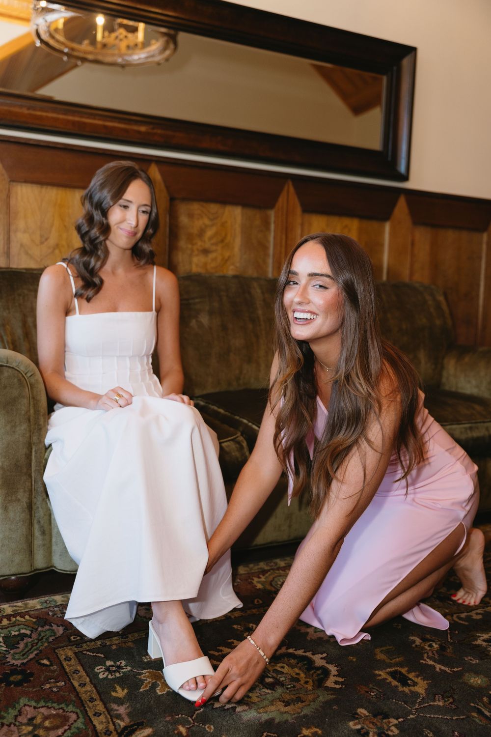 Bridesmaid helping bride put on white heels while sitting on a couch before the wedding.