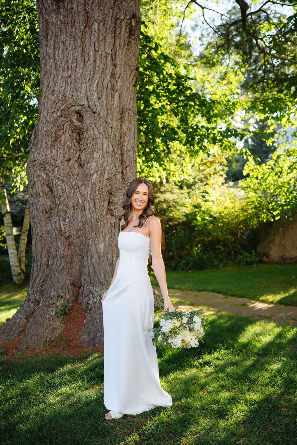 Bride in elegant white gown holds bouquet while standing beside large tree in sunlit garden.