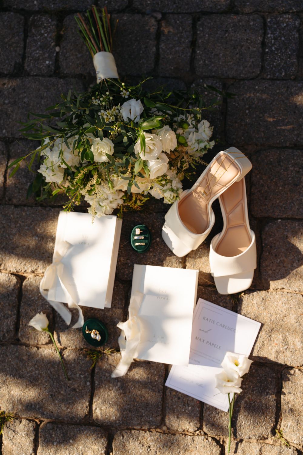 Elegant wedding flat lay featuring nude heels, white floral bouquet, green ring boxes, and stationery on cobblestone.
