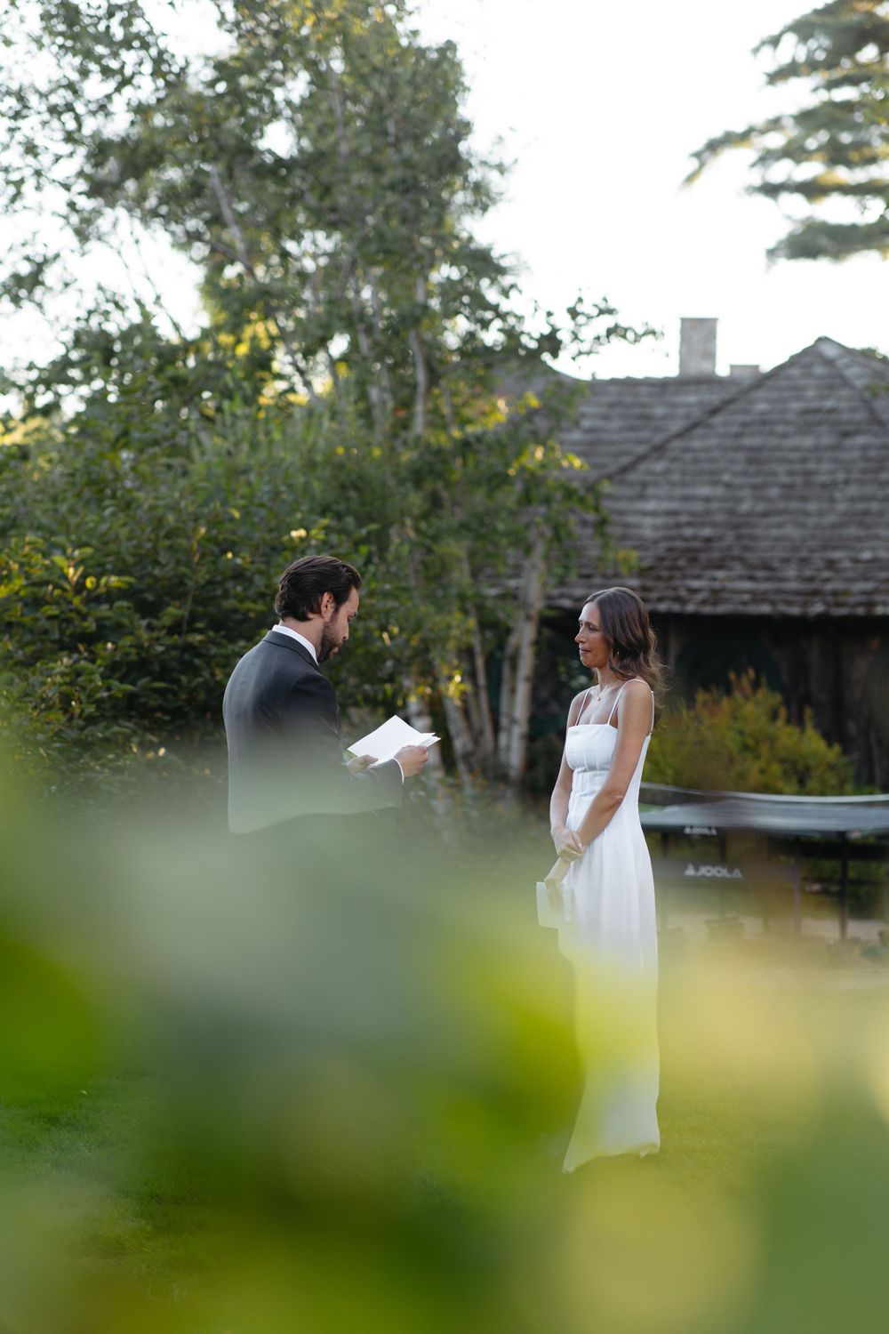 Groom reads vows to smiling bride during intimate outdoor wedding ceremony near rustic building.