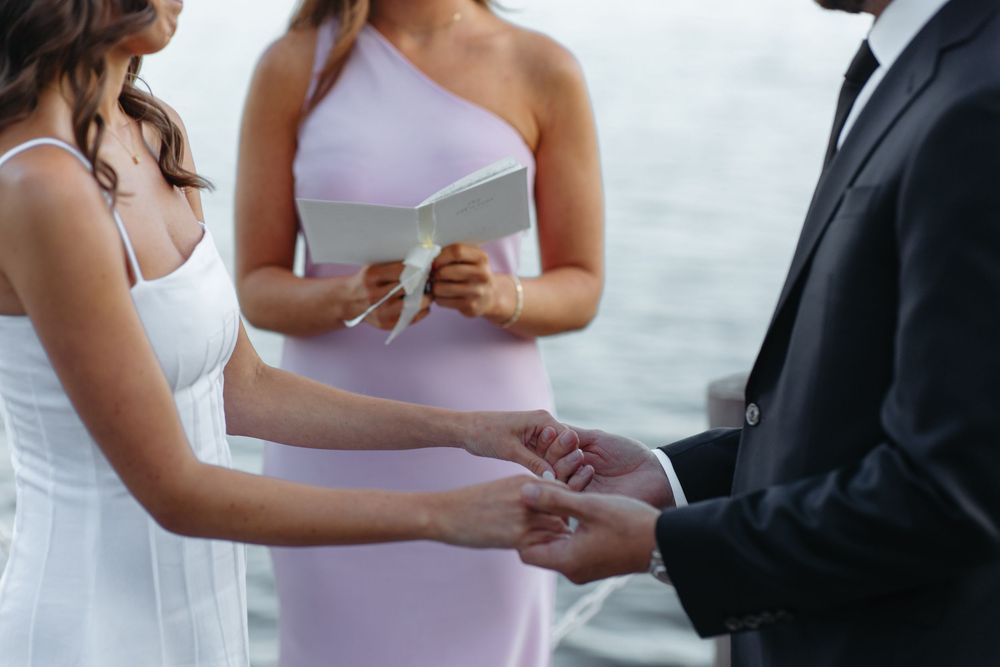 Bride places wedding ring on grooms hand as officiant in pink dress reads ceremony.