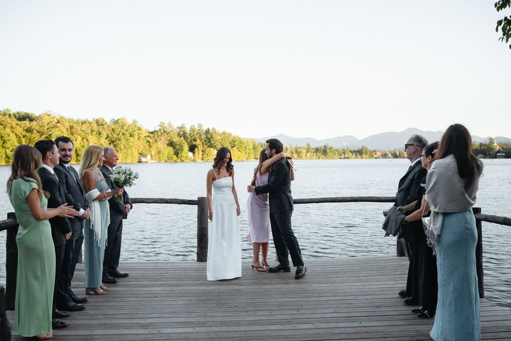Intimate lakeside wedding ceremony on wooden dock with small group of guests at sunset.