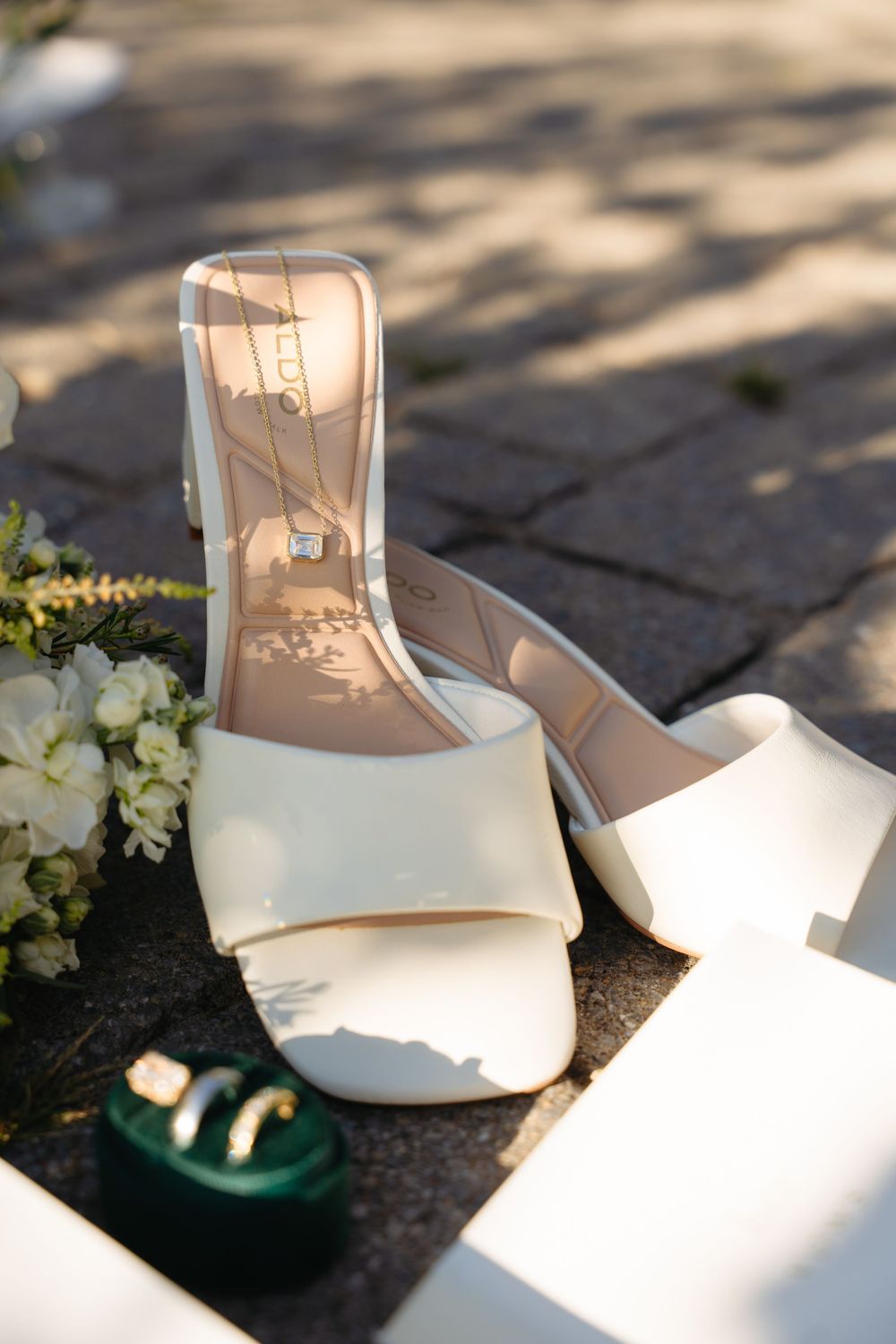 White bridal heels with gold necklace and green ring box on stone pavers at outdoor wedding.