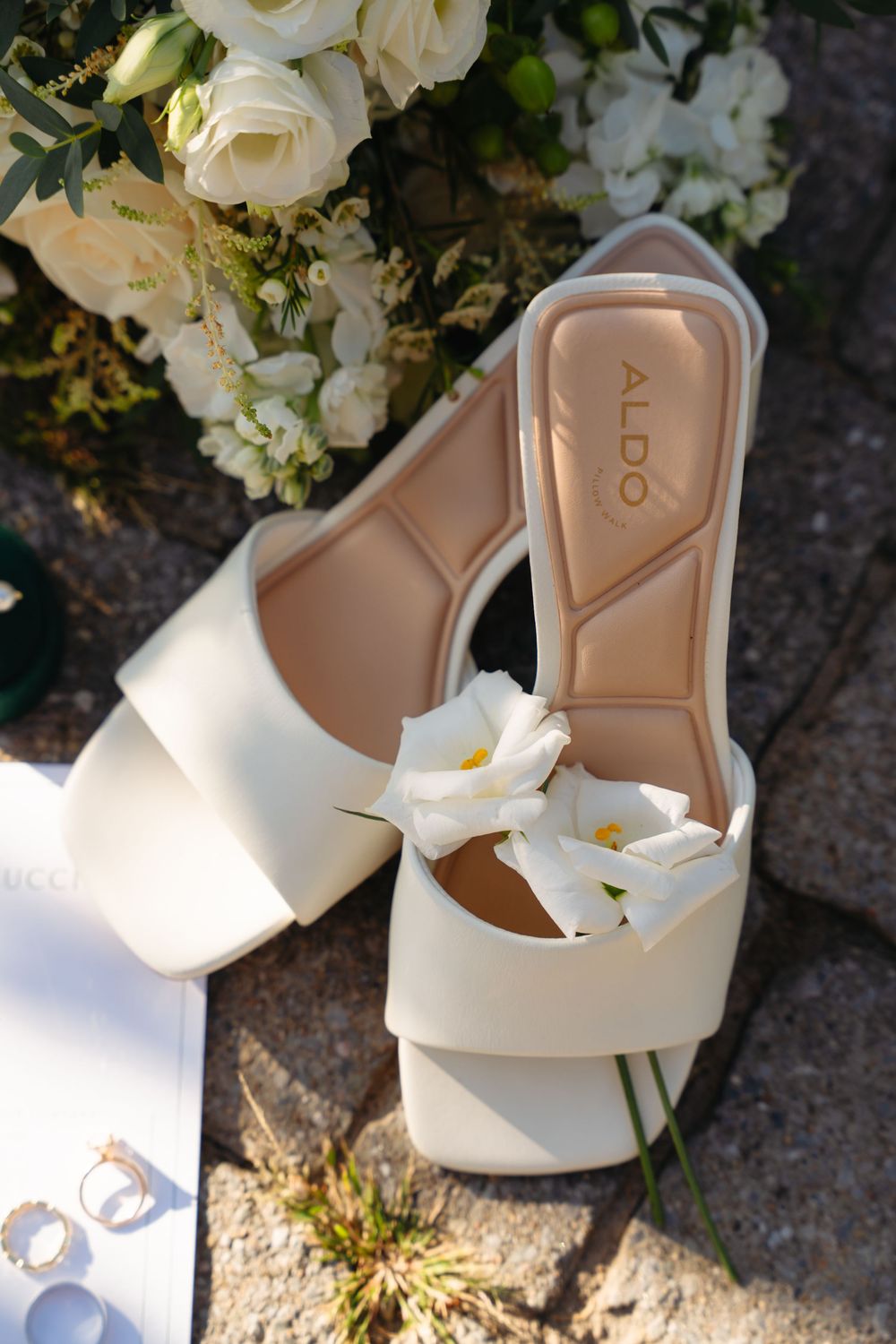 White ALDO bridal heels with floral detail beside wedding rings and white rose bouquet on stone surface.