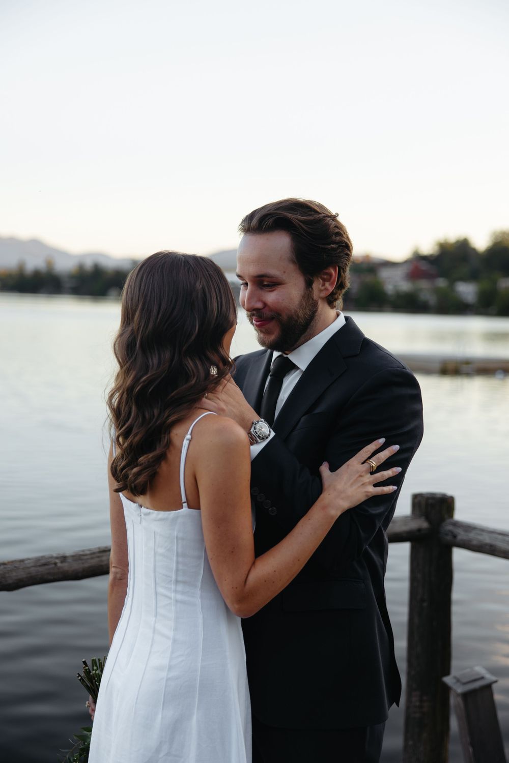 A smiling groom in a dark suit embraces his bride in a white dress by a lakeside at dusk.