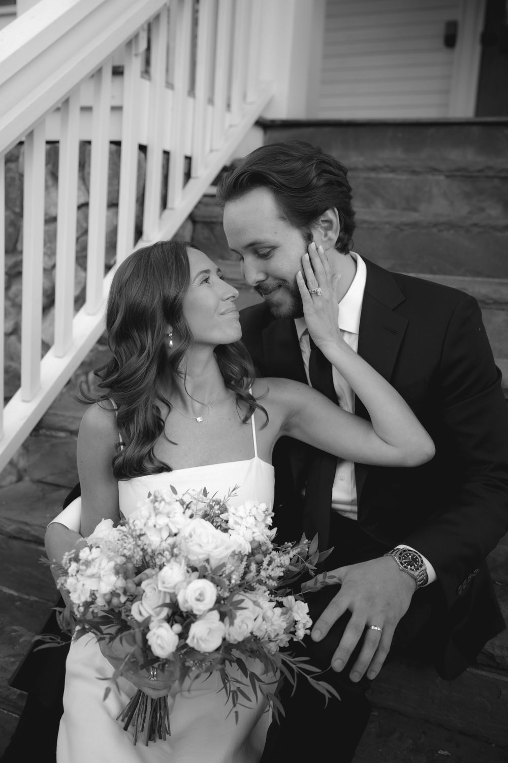 Black and white photo of bride and groom sharing tender moment on steps, bride holding floral bouquet.