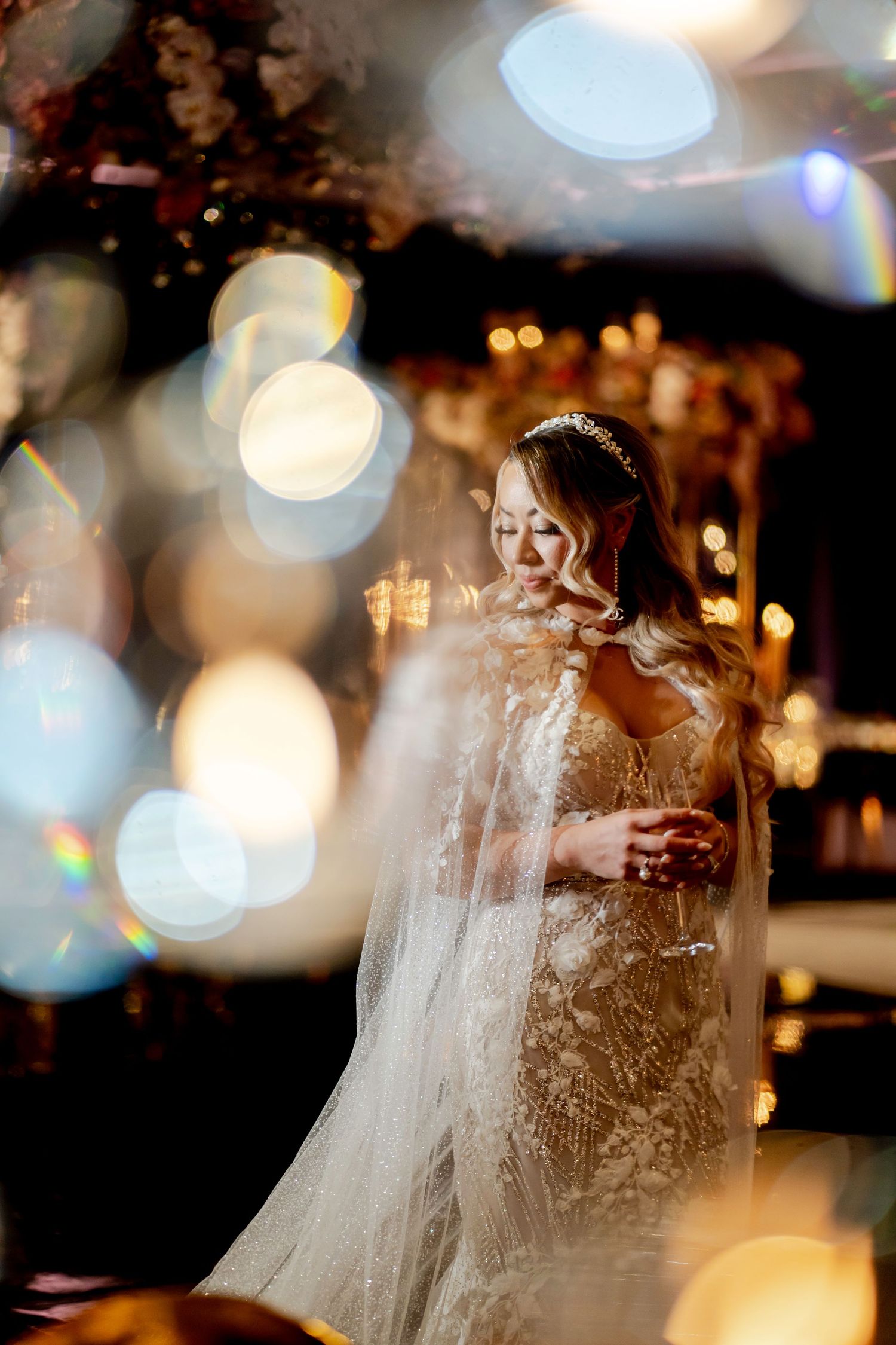 Elegant bride in a sparkling lace wedding gown with a sheer cape and tiara, glowing amid golden bokeh lights at a luxurious reception.