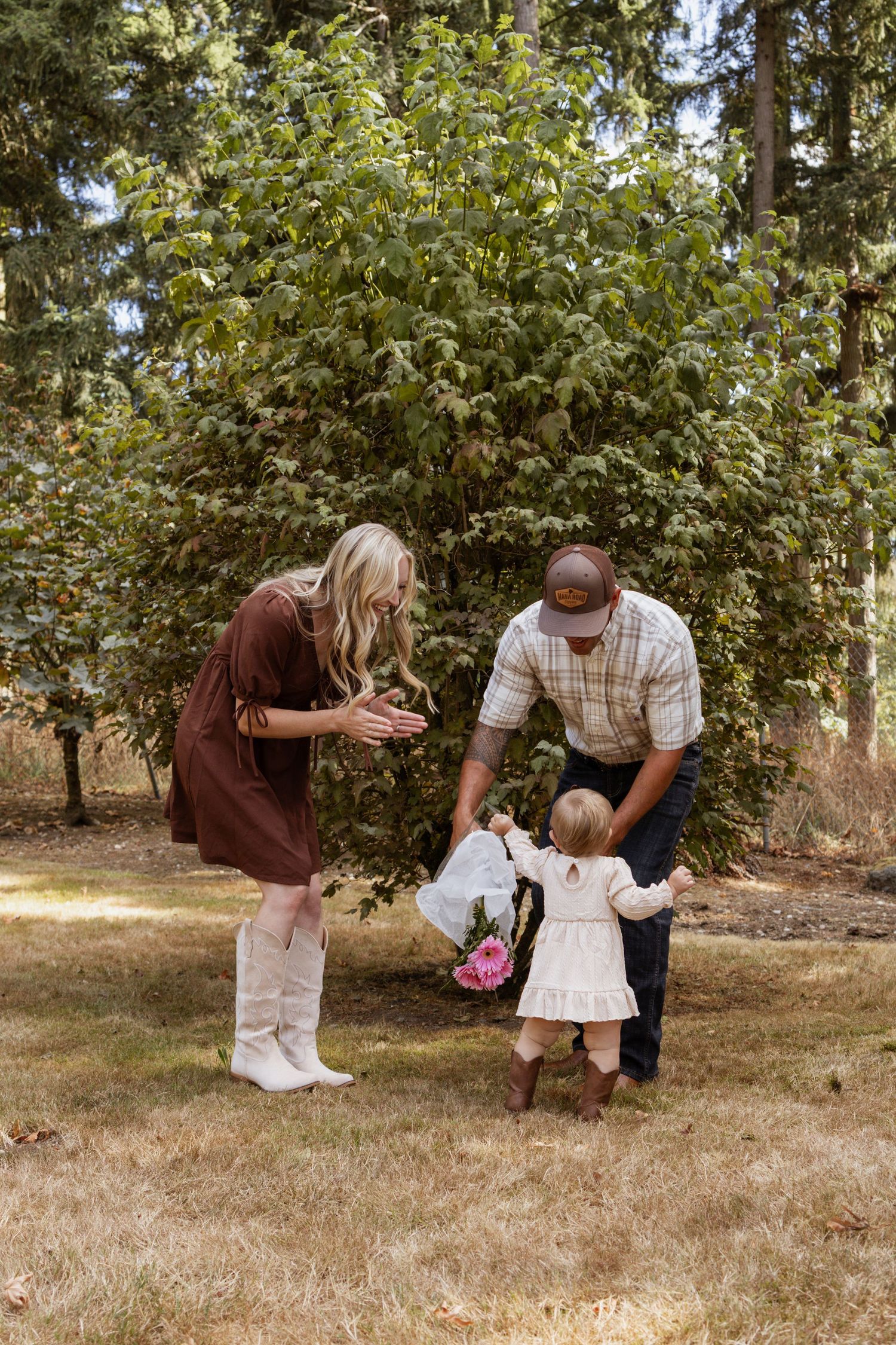 A young family with parents in fall attire helping their toddler walk on a grassy lawn surrounded by green trees.
