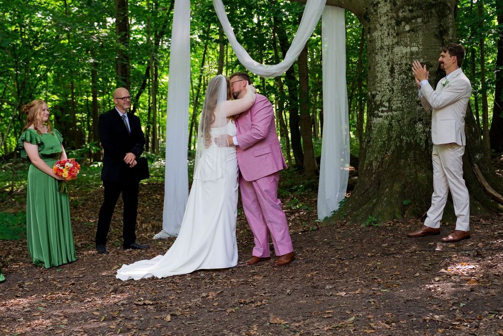Classic bride in white and groom in a pink suit share a first kiss during an outdoor forest wedding ceremony at The Centennial Barn.