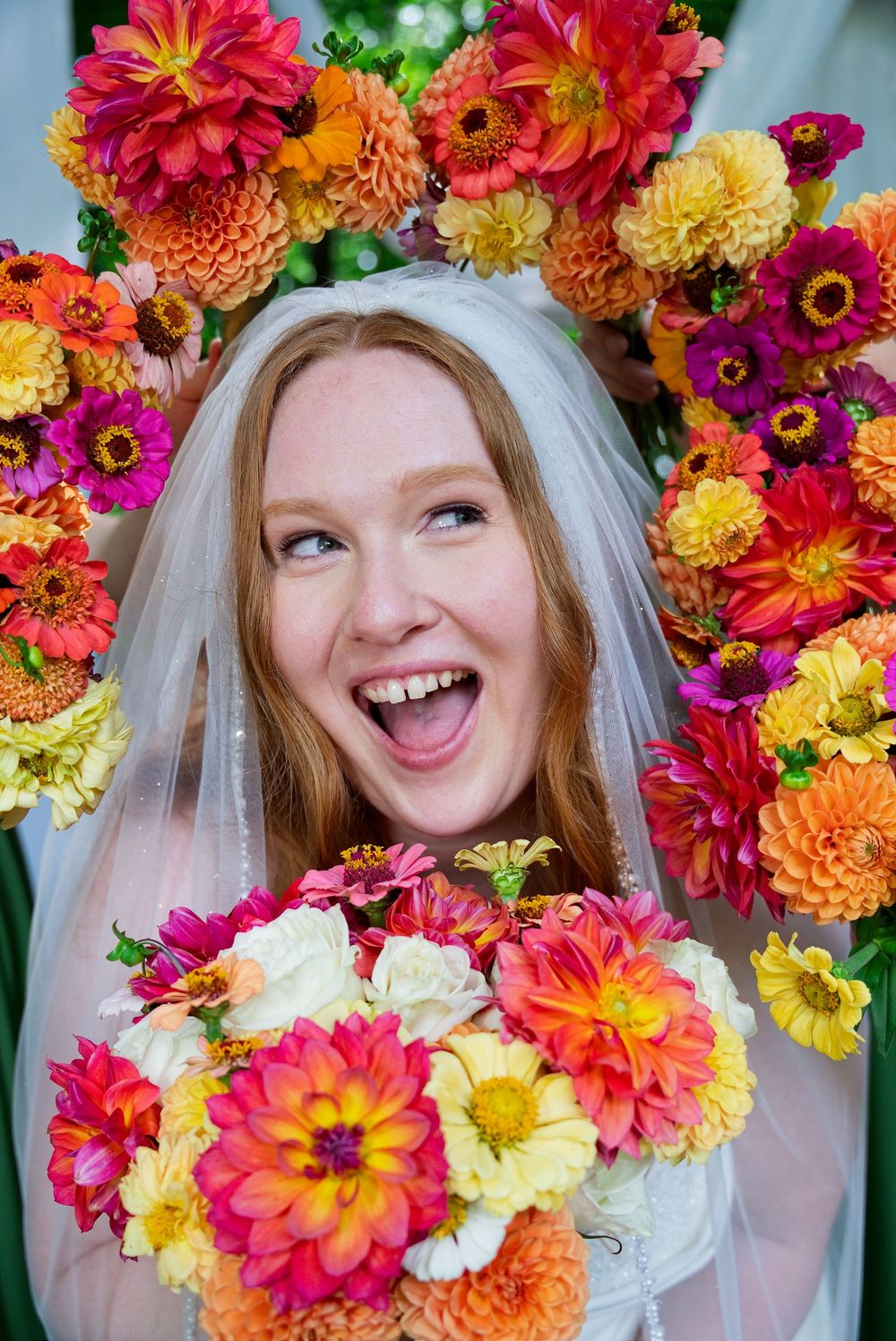 A smiling bride with a veil holds a vibrant bouquet of homegrown pink zinnias, orange dahlias, and cream garden roses.