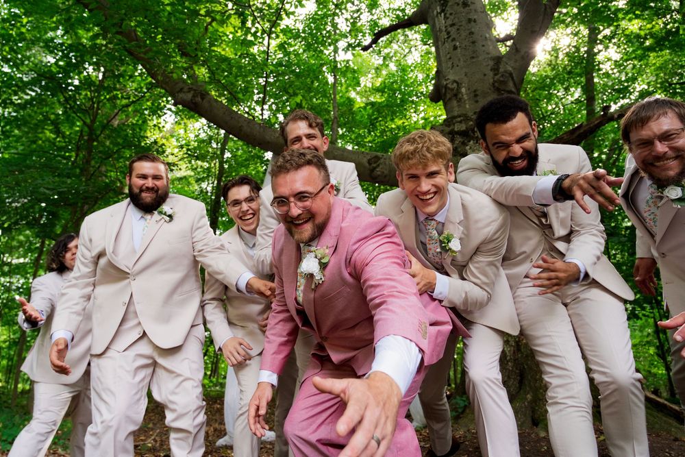 A groom in a pink suit laughs during fun, high-energy wedding portraits with his groomsmen in the woods.