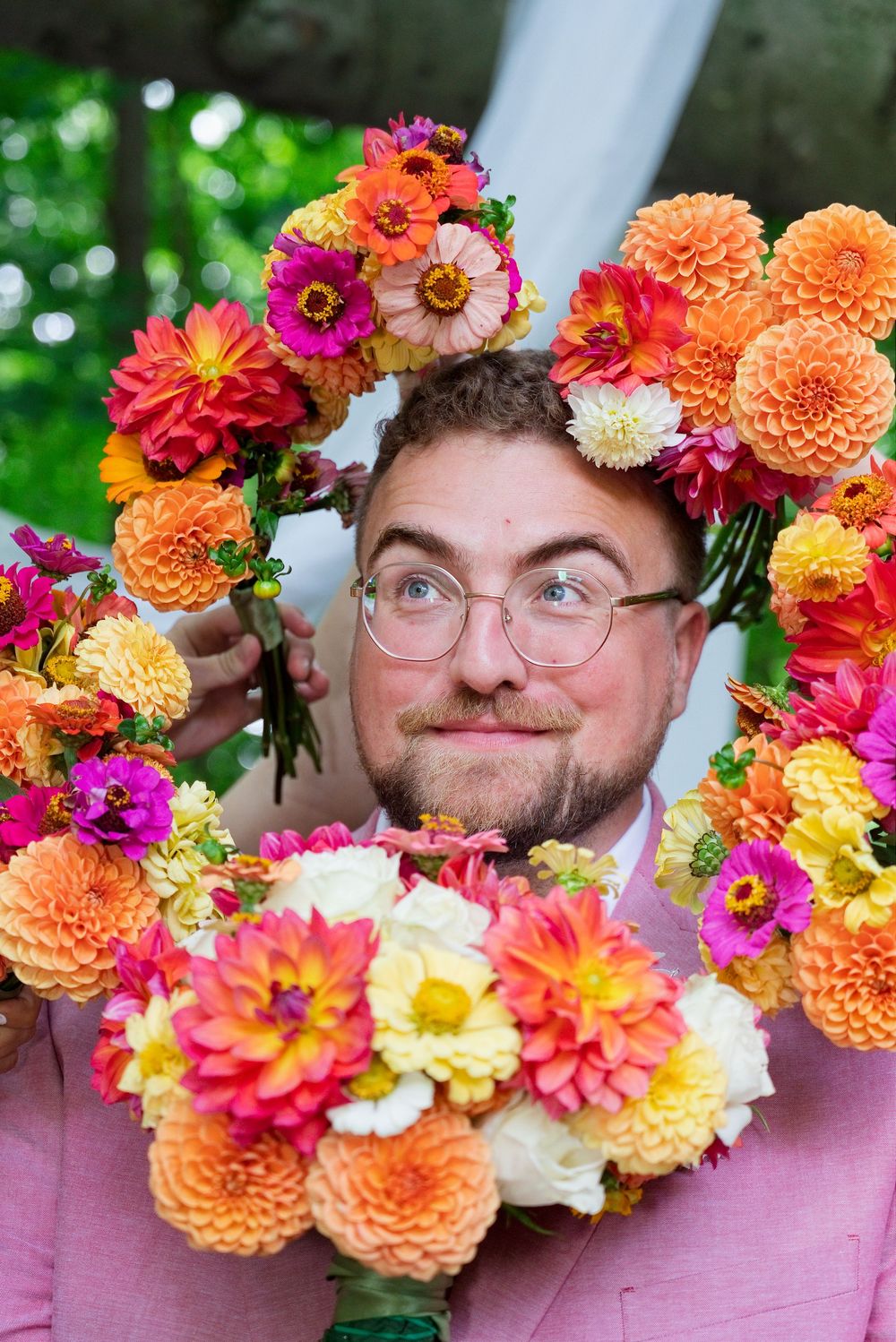 Vibrant bouquets of homegrown pink zinnias, orange dahlias, and cream garden roses surround a bemused groom.