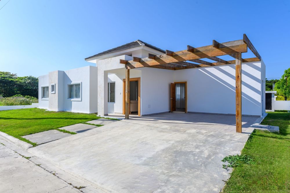 Modern white single-story home with wooden pergola, concrete driveway, and lush green lawn under a clear blue sky.