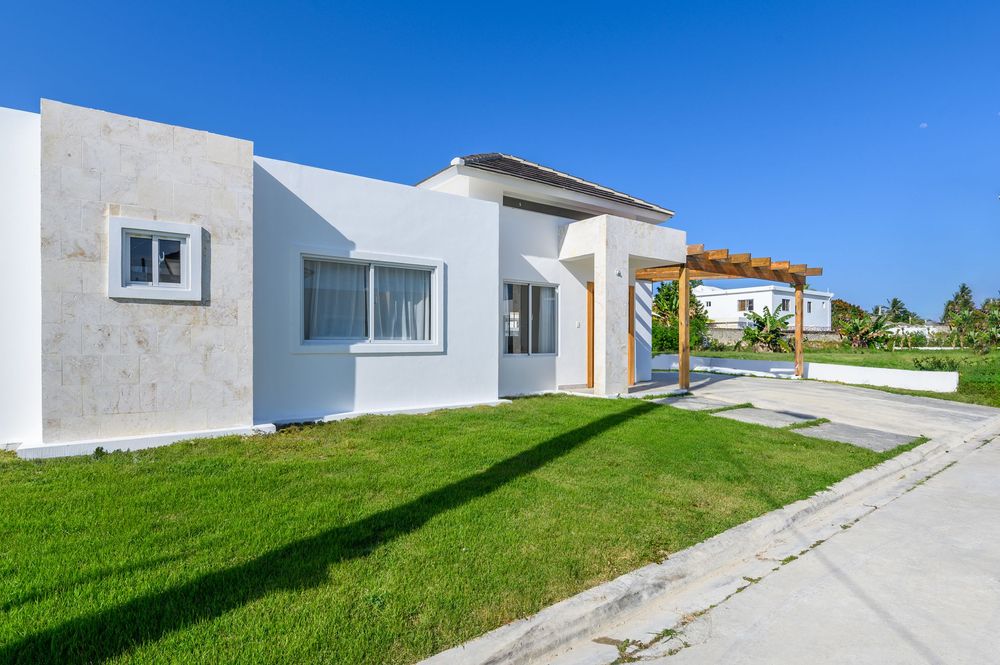 Modern white stucco beach house with lush green lawn, pergola entrance, and clear blue sky in tropical setting.