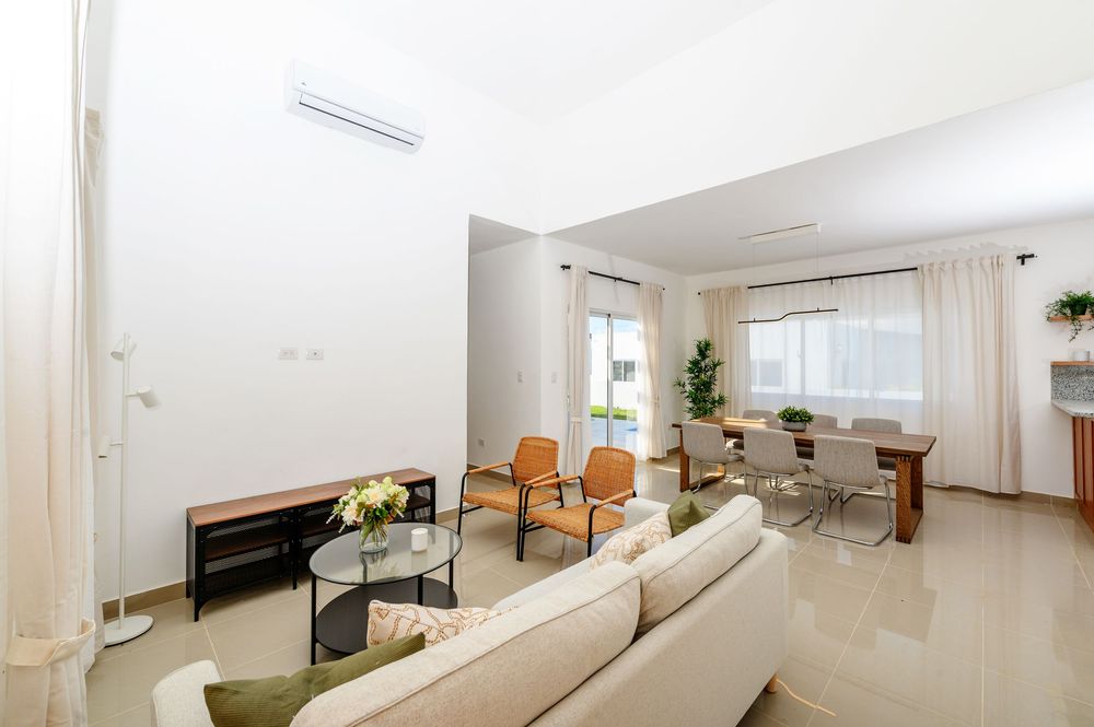 Bright modern living room with beige sofa, glass coffee table, dining area, and white walls with AC unit.