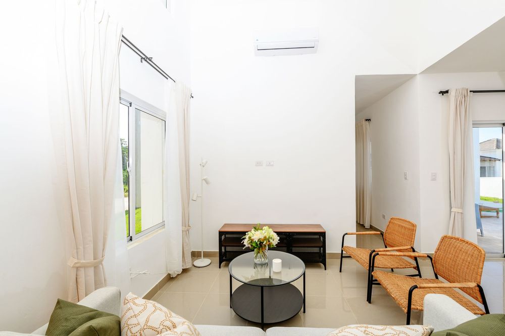 Bright modern living room with white walls, rattan chairs, round coffee table, green sofa, and mini-split AC unit.