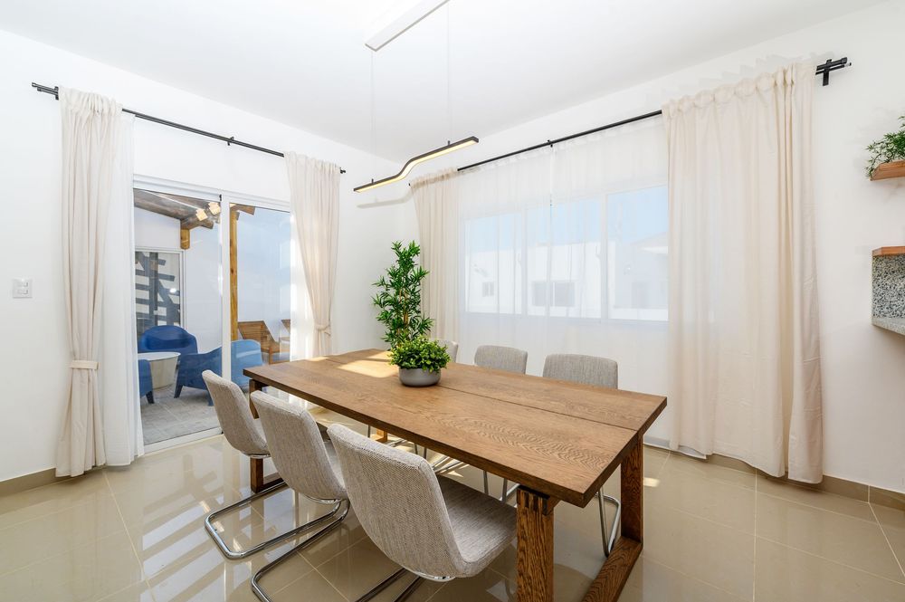 Modern dining room with wooden table, white chairs, pendant light, and bright windows with sheer curtains.