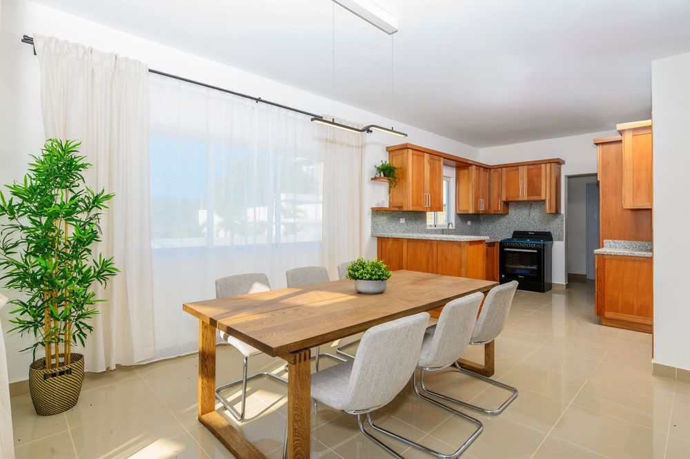Bright dining area with wooden table, white chairs, and open kitchen featuring cherry cabinets and large windows.