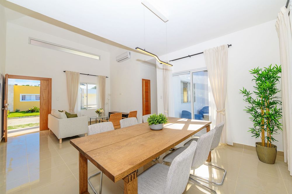 Bright modern dining room with wooden table, white chairs, pendant light, and large windows overlooking a sunny patio.