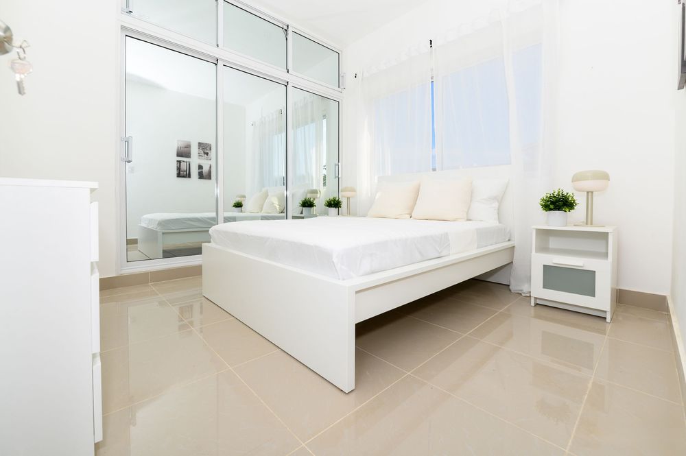 Bright white minimalist bedroom with mirrored wardrobe, white bed frame, nightstand, and large window flooding the room with natural light.