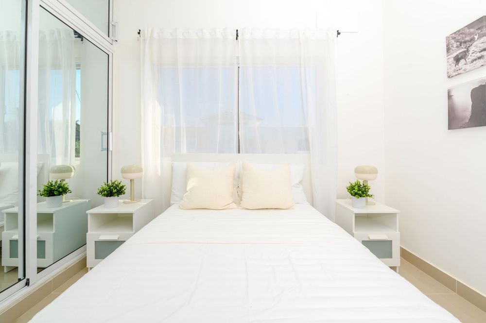 Bright white bedroom with a double bed, white bedside tables with green plants, and sheer curtains by the window.