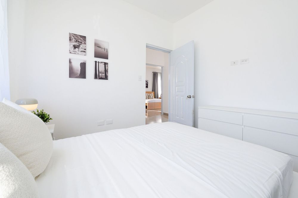 Bright white minimalist bedroom with a large bed, dresser, and black-and-white wall art, leading to an open doorway.