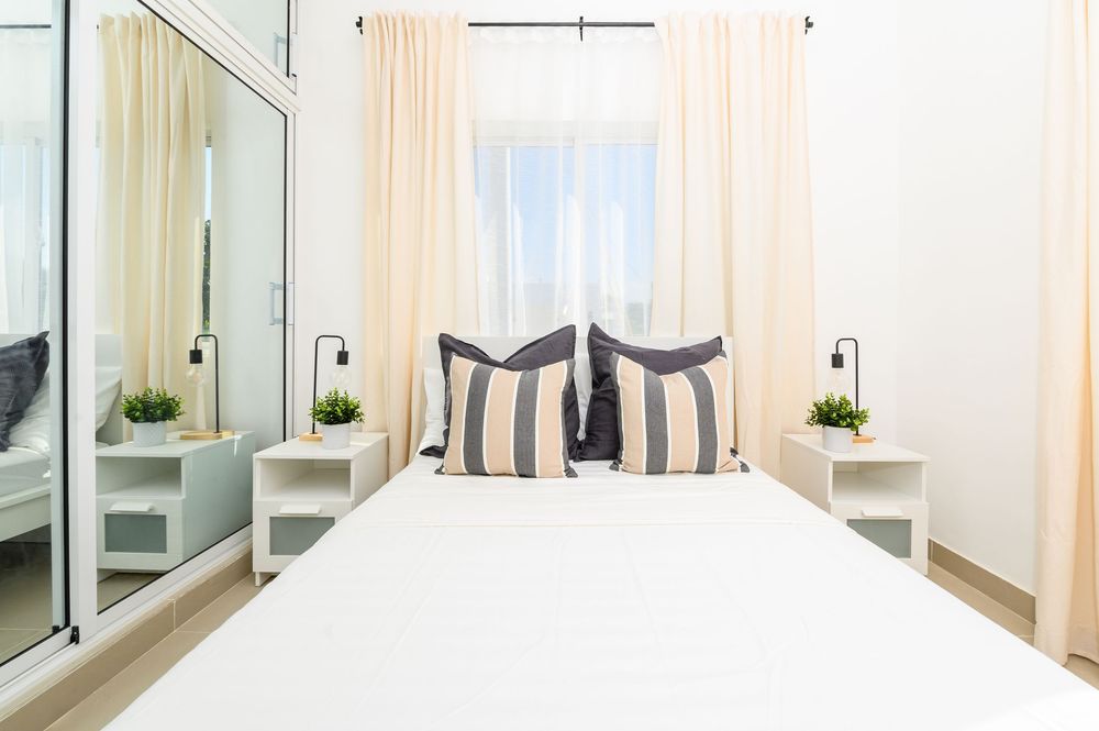 Bright white bedroom with striped pillows, mirrored wardrobe, twin nightstands with plants, and sheer curtains.