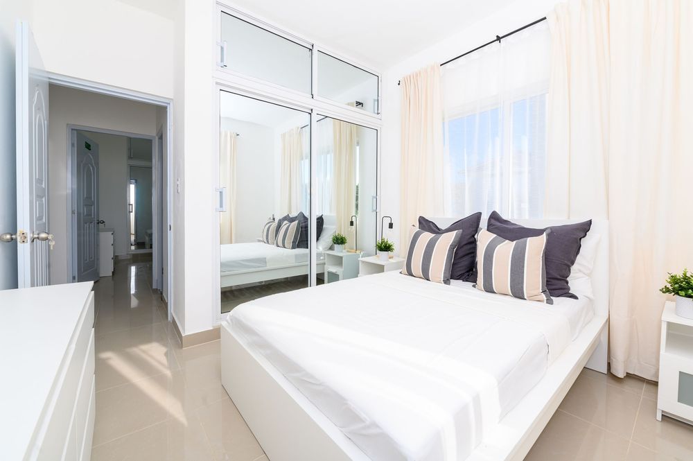 Bright white bedroom with mirrored wardrobe, striped accent pillows, and sheer curtains in a modern minimalist design.
