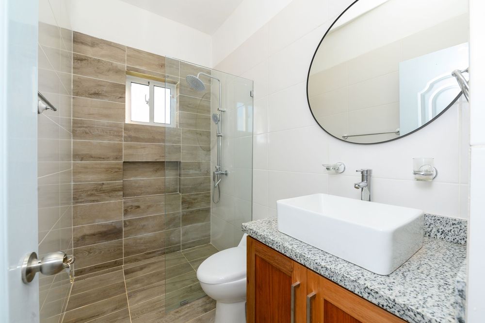Modern bathroom with walk-in tile shower, vessel sink on wood vanity, round mirror, and granite countertop.