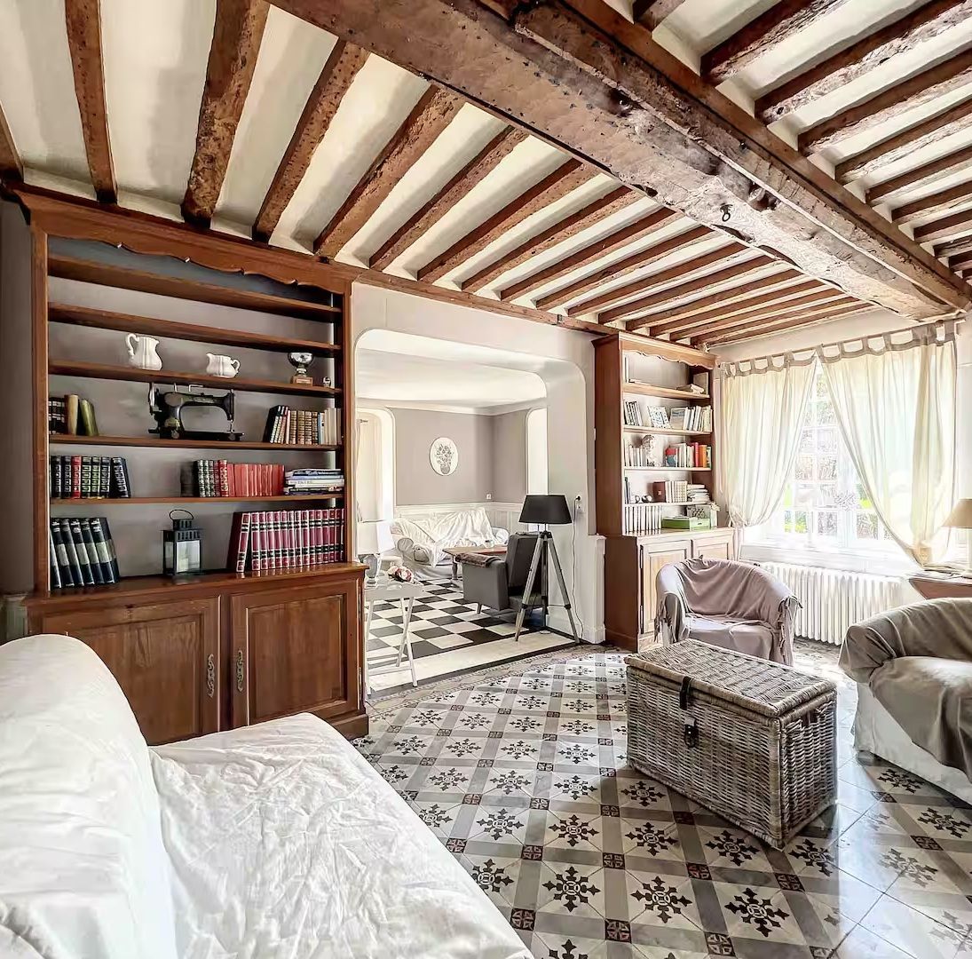 Elegant French living room with exposed wooden beams, patterned tile floors, built-in bookshelves, and white sofas.