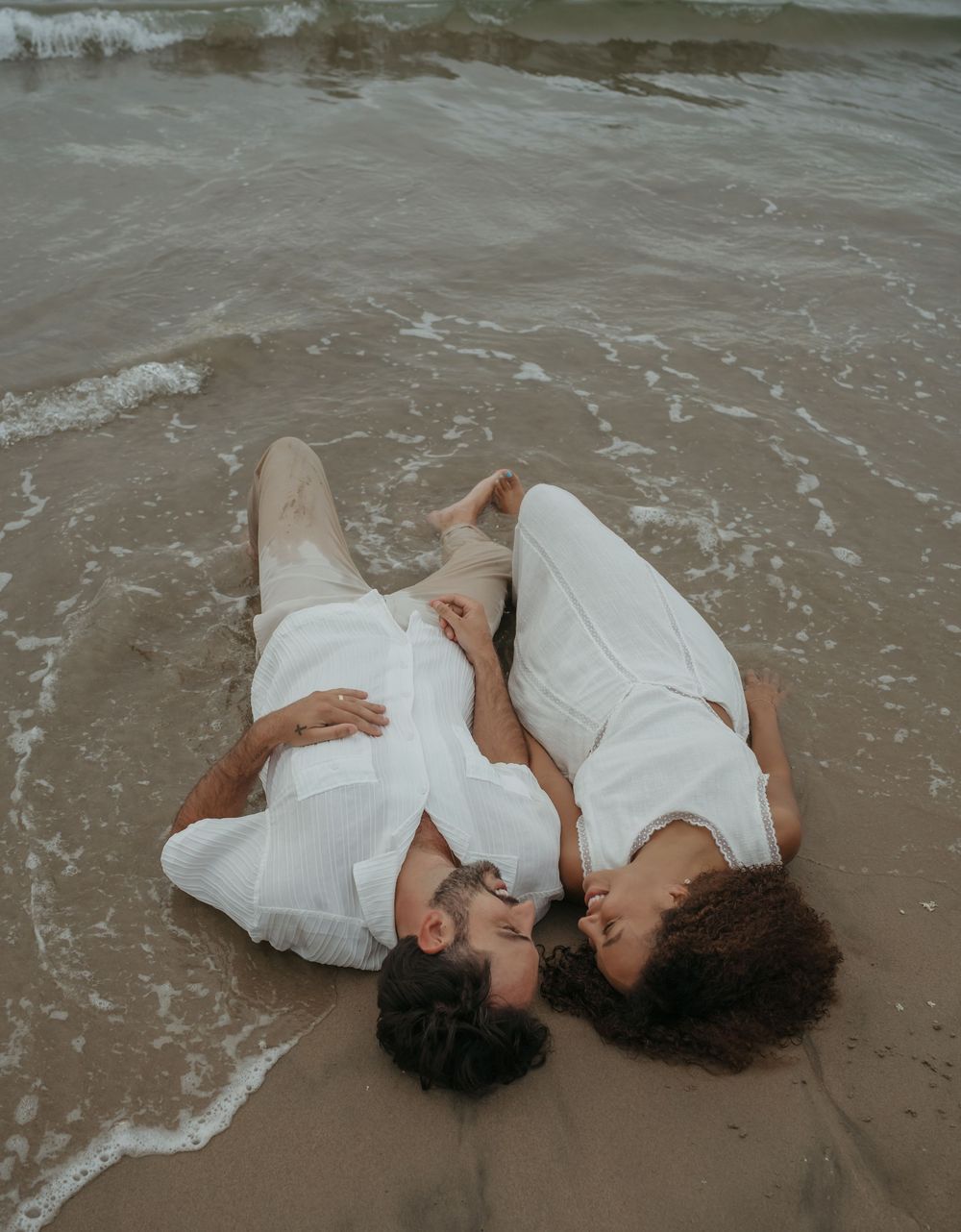 Couple in white clothing lying on sandy beach shore, sharing a romantic kiss as ocean waves wash around them.