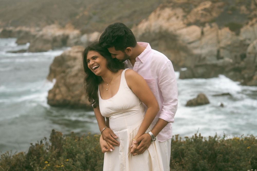Couple sharing a joyful moment by the ocean cliffs, woman in white dress laughing as man embraces her.