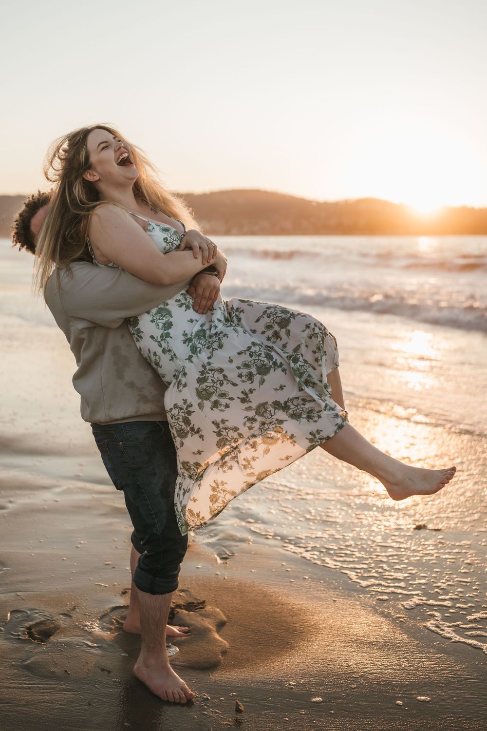 Couple laughing joyfully on a sandy beach at golden sunset, man lifting woman in floral dress by the ocean waves.