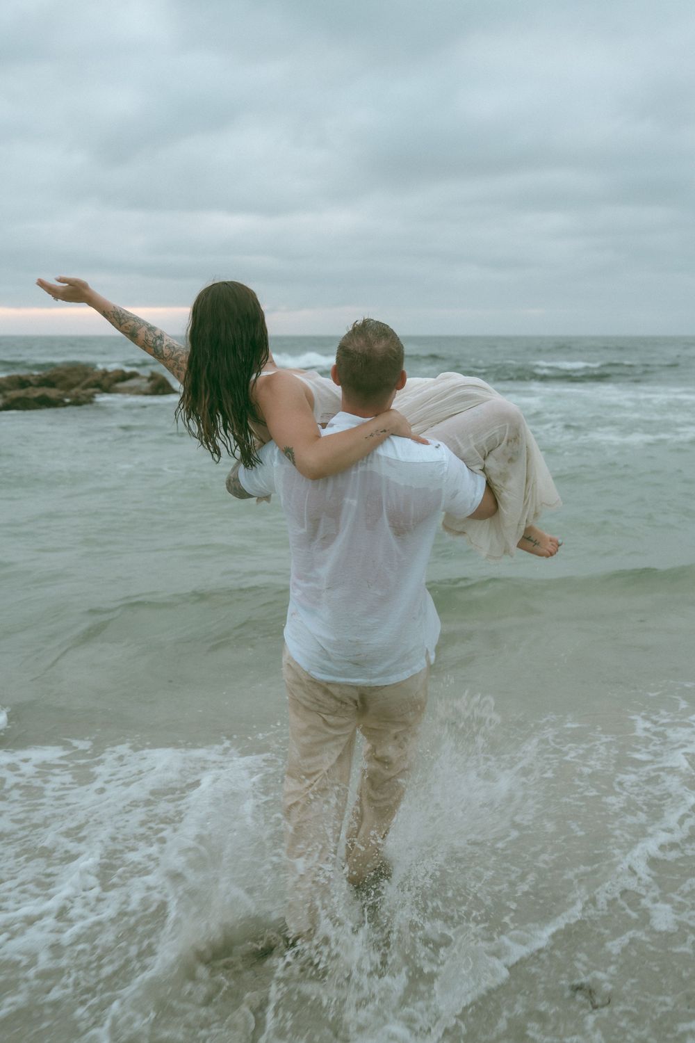A man carries a woman in his arms while wading through ocean waves on a cloudy beach day.