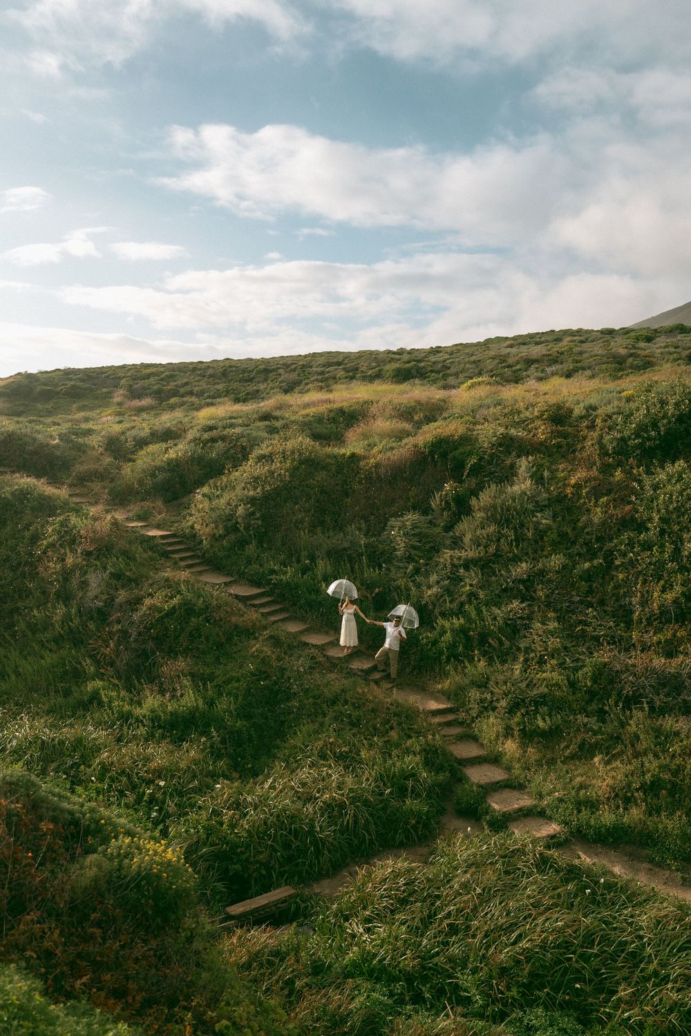 Two figures in white dresses carrying parasols walk up stone steps through lush green hillside under a cloudy sky.