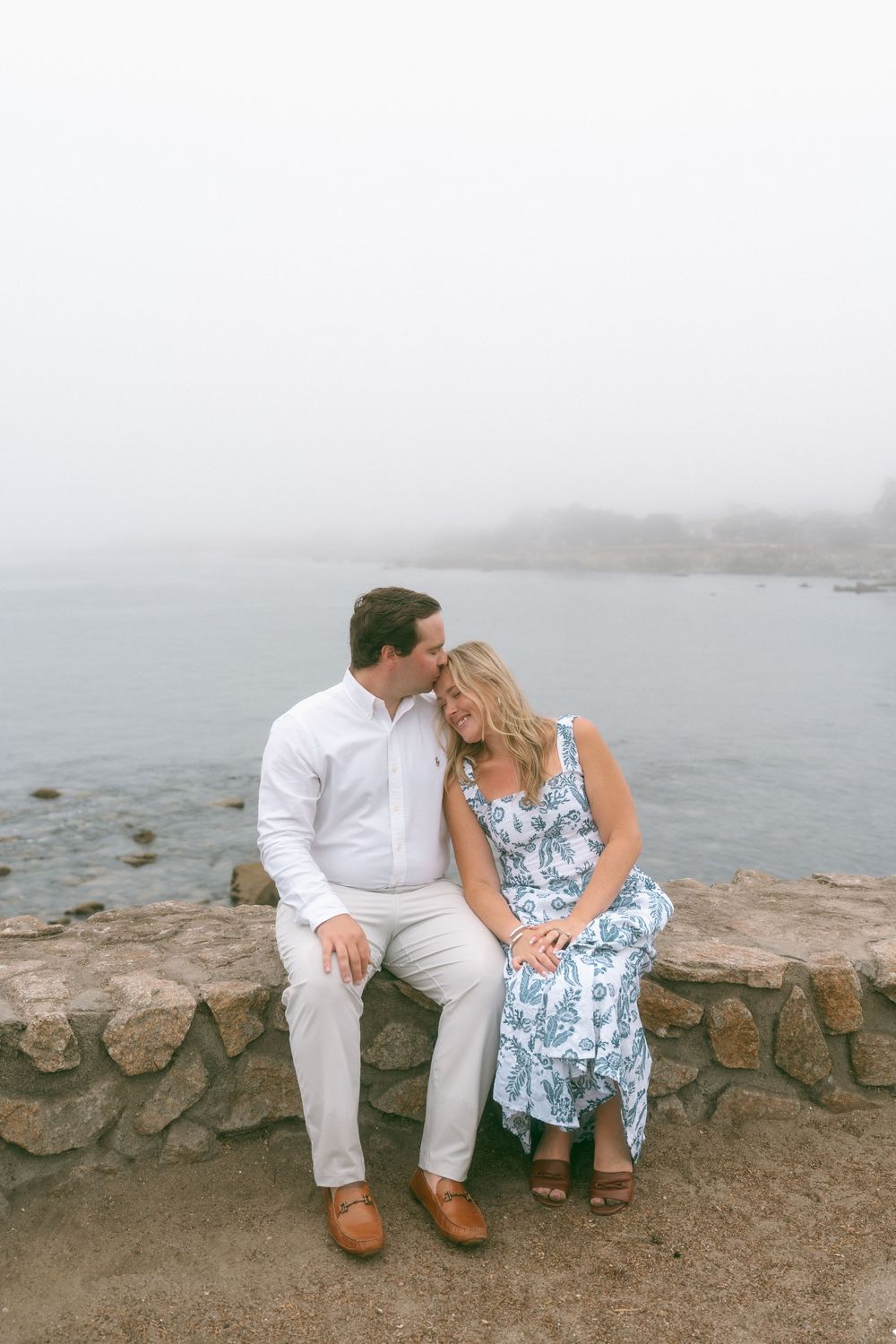 Couple sitting on stone wall by foggy ocean, man in white shirt kissing woman’s forehead, blue floral dress.