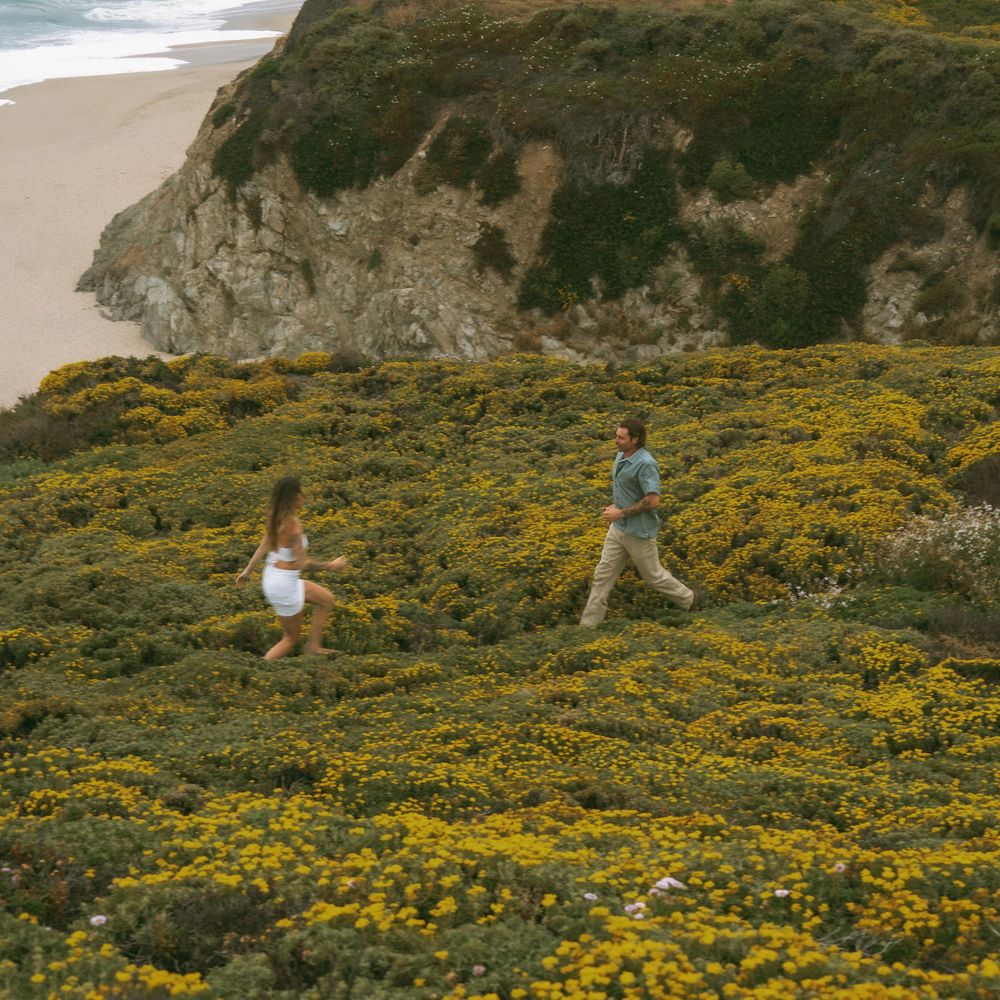 Two people running through vibrant yellow wildflowers on a coastal hillside with sandy beach and rocky cliffs in background.