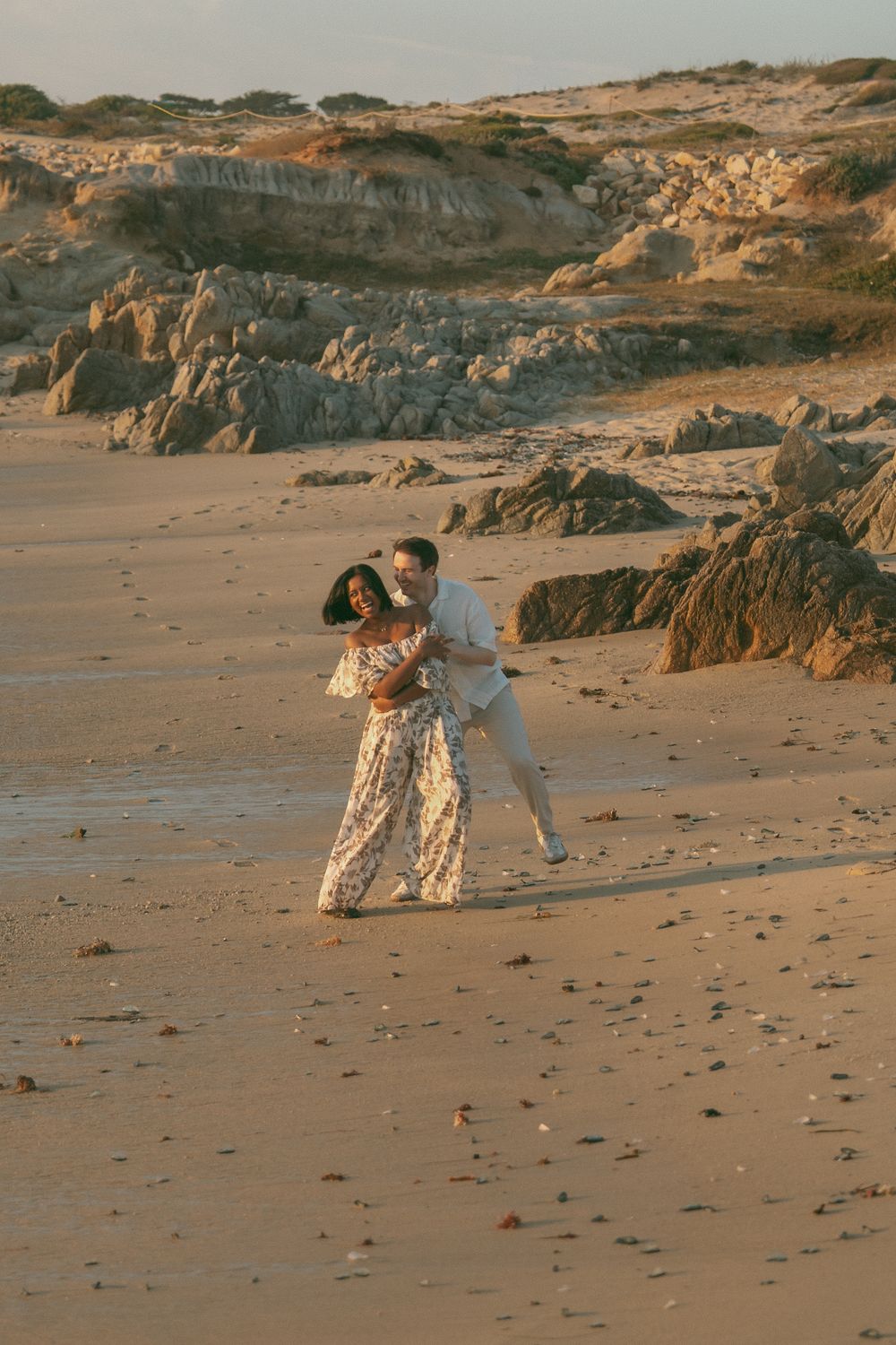 Couple dancing romantically on a sandy beach at golden hour with rocky cliffs in the background.