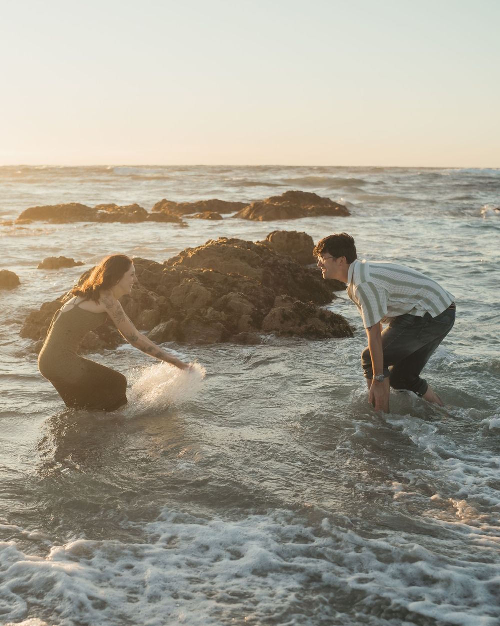 Two people crouching in ocean waves near rocky shore at golden hour sunset.