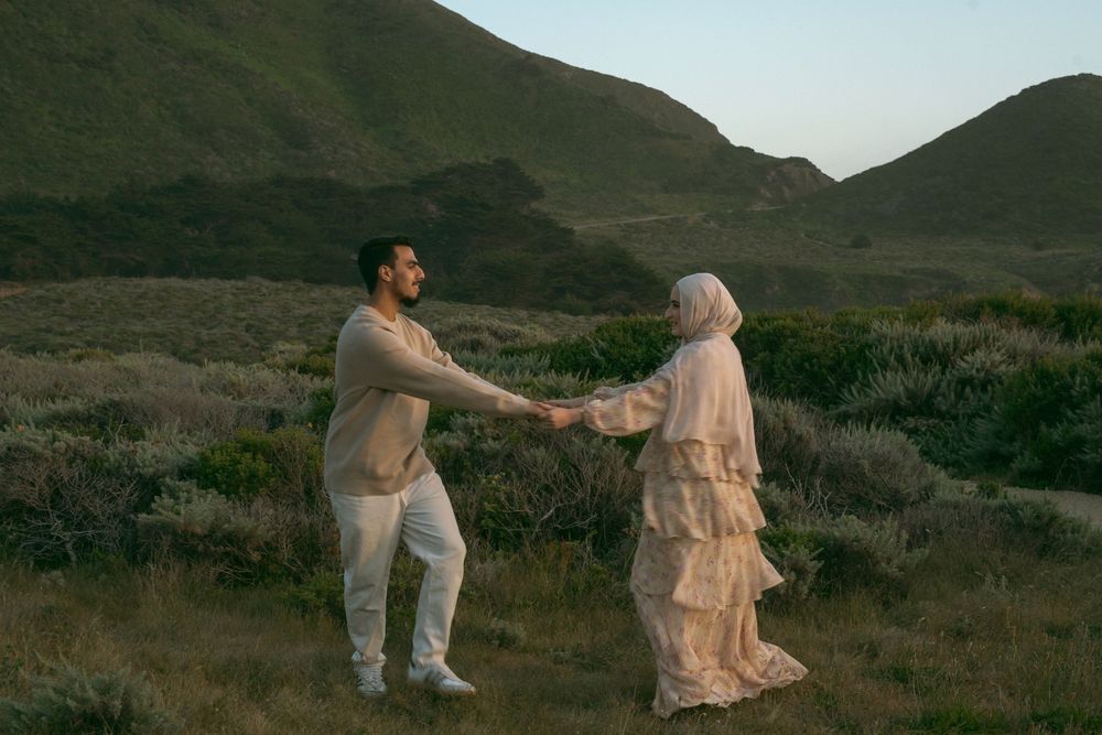 Couple in white attire holding hands in a scenic green hillside landscape at dusk.