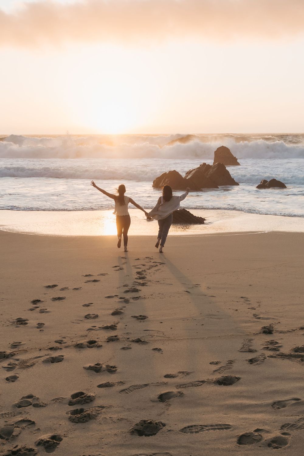 Two women holding hands running toward sunset waves on a sandy beach with rocky outcrops and footprints in the sand.