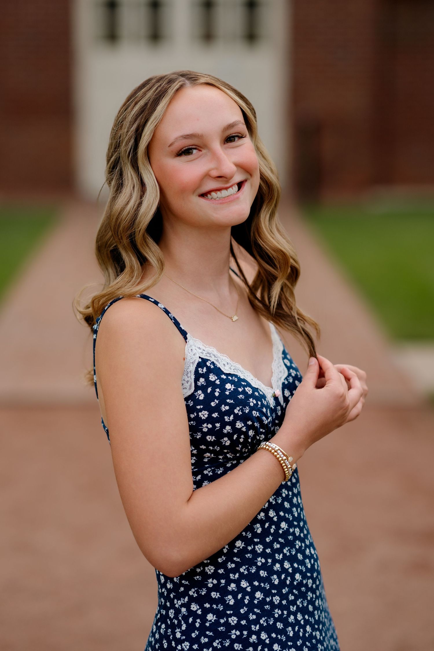 Young woman smiling in navy floral sundress with lace trim, posing outdoors near brick building for senior portrait.