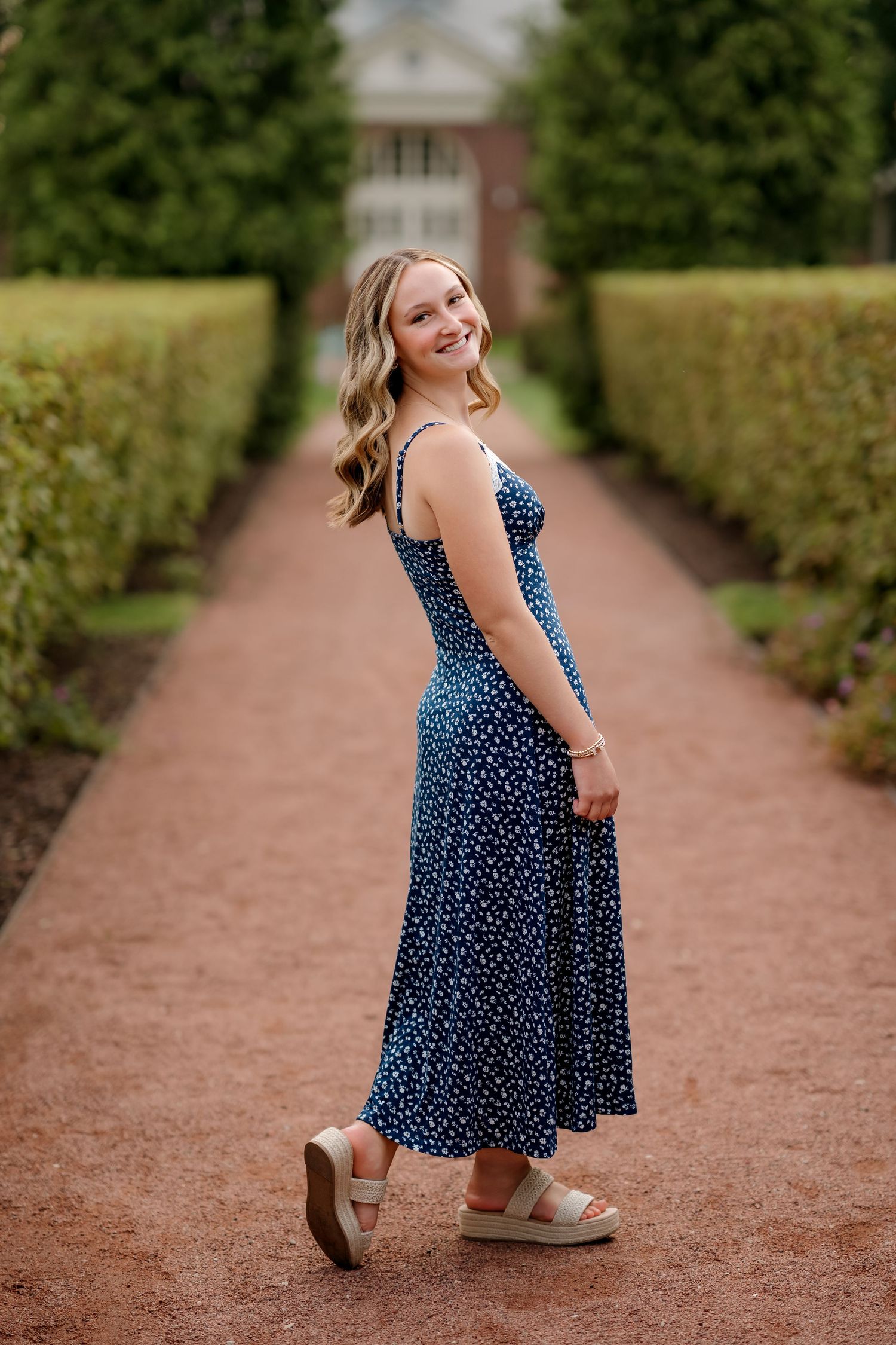 Young woman in navy floral midi dress poses on brick garden path flanked by tall hedges.