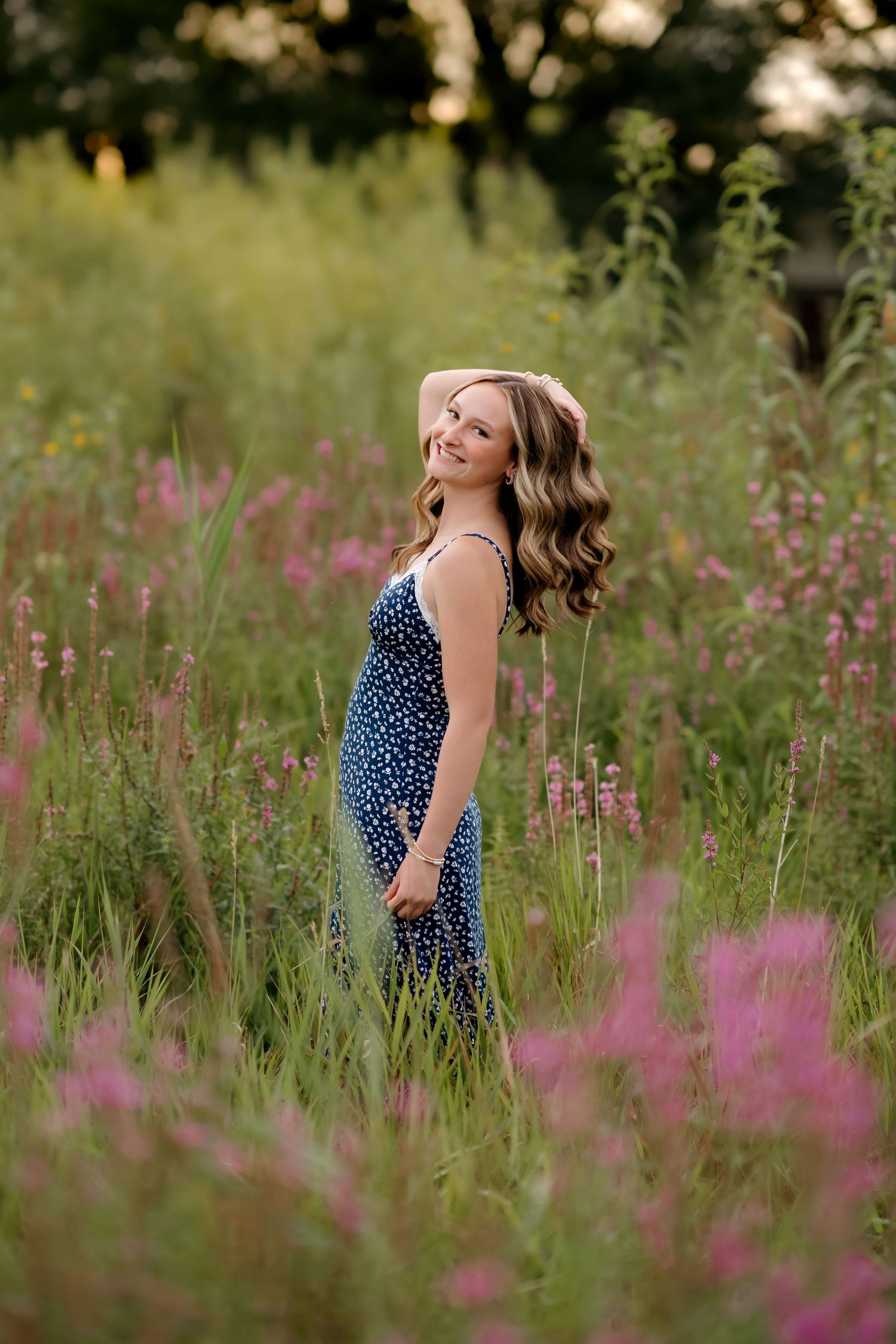 Young woman in blue floral dress smiling in a lush wildflower meadow with pink blooms at golden hour.