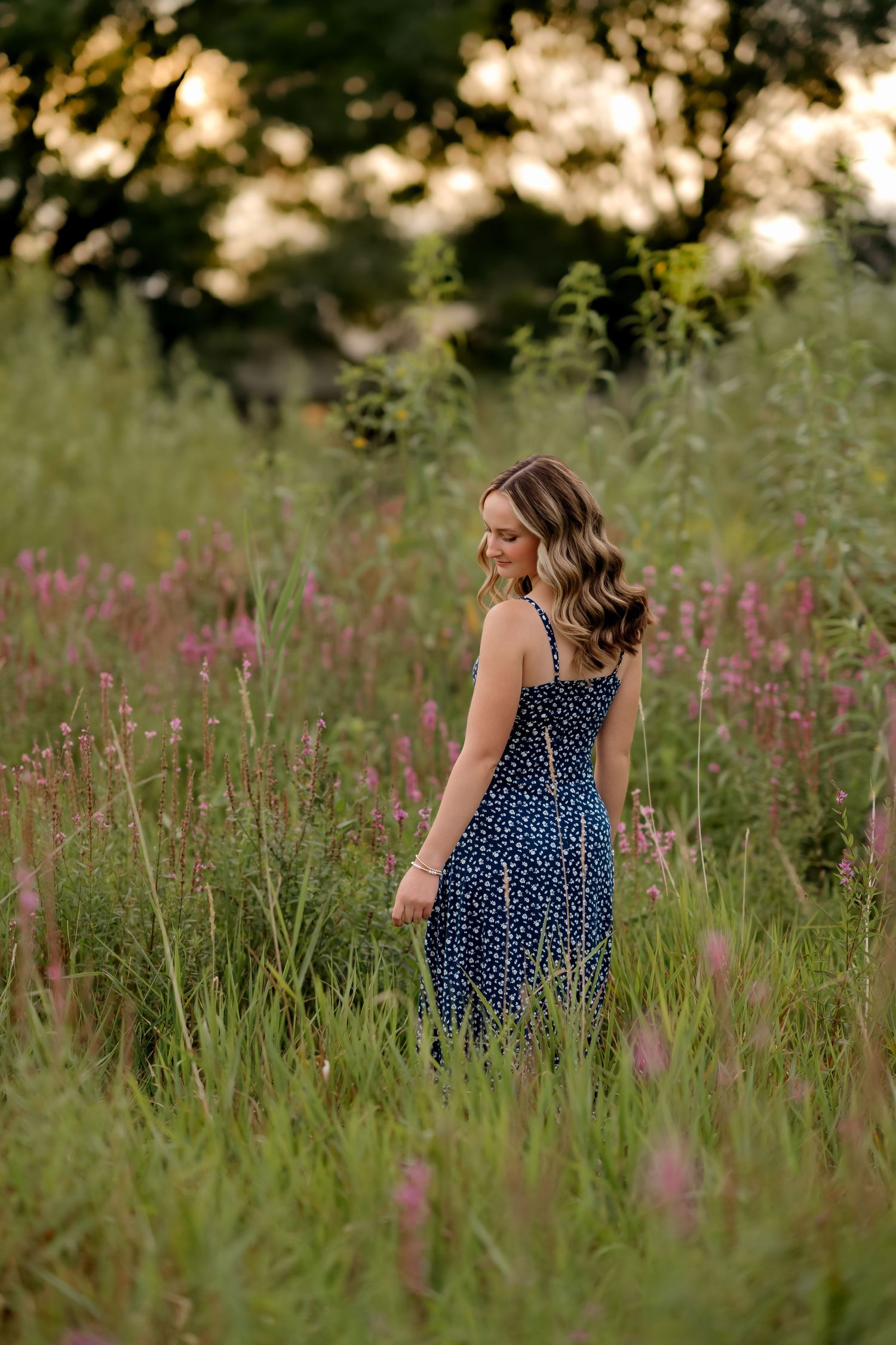 Young woman in blue floral sundress standing among pink wildflowers in a lush green field at golden hour.