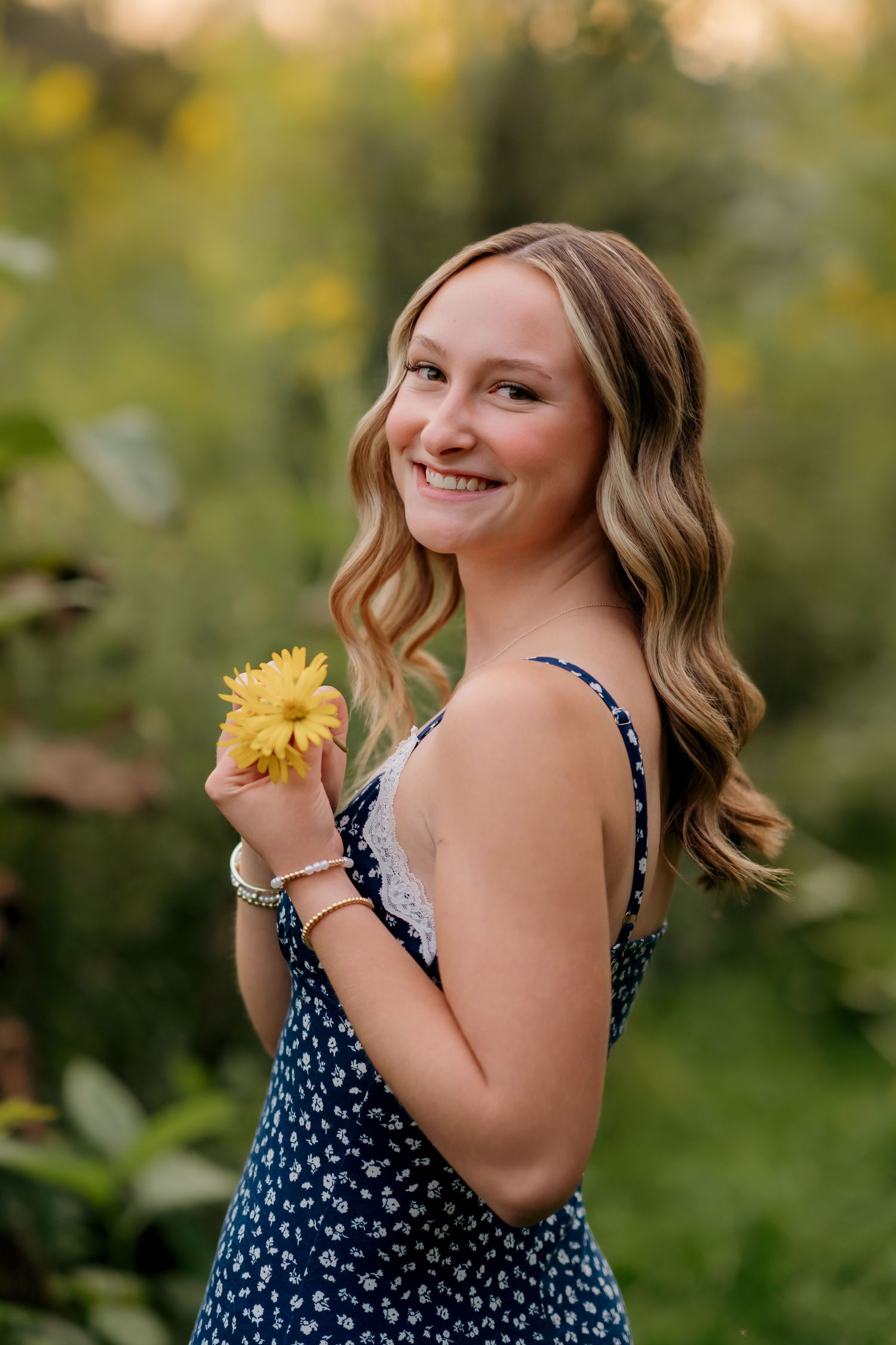 Smiling young woman in blue floral dress holding a yellow daisy flower outdoors in a lush green garden setting.