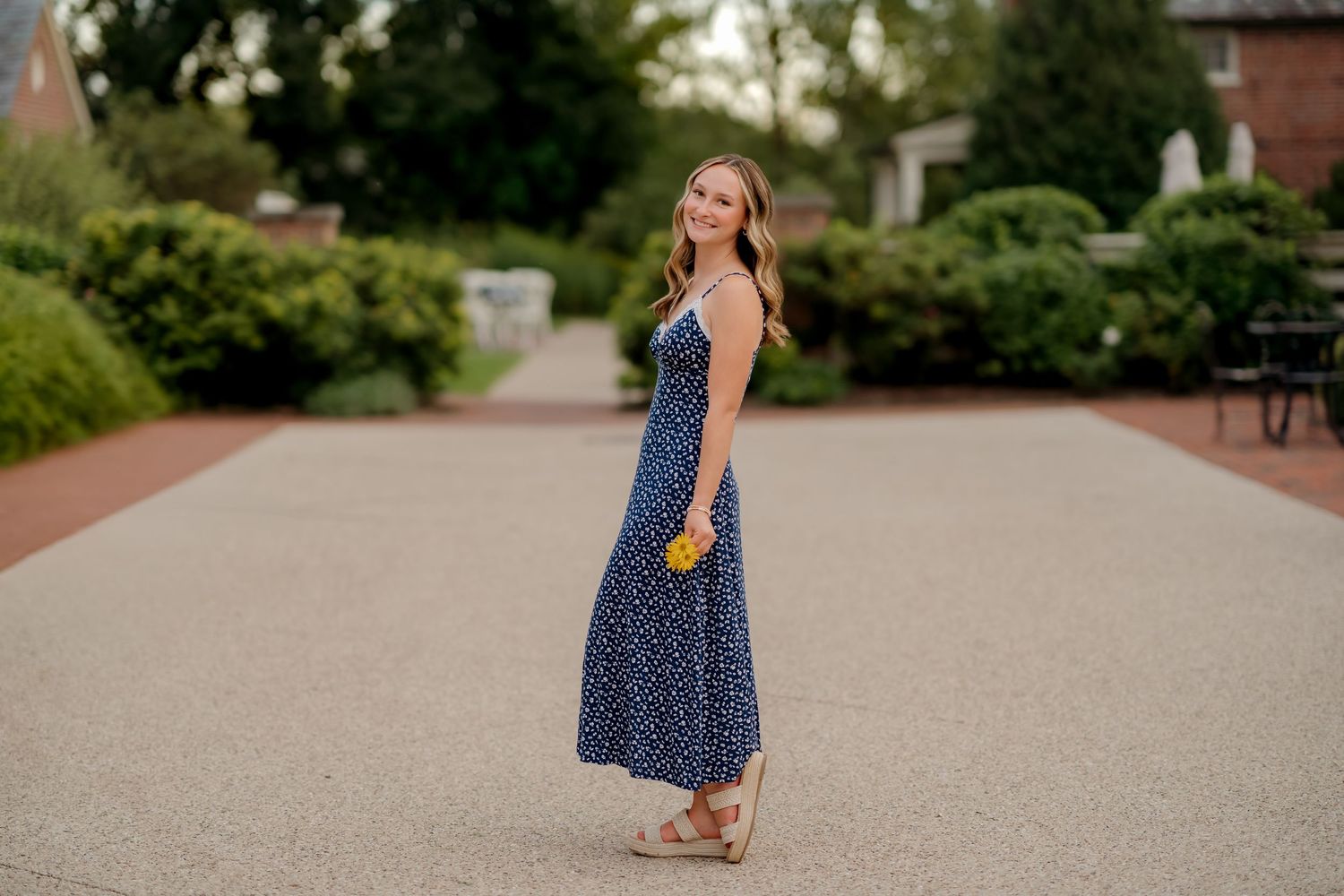 Young woman in navy blue floral maxi dress holding yellow flowers poses on a garden path with lush greenery.