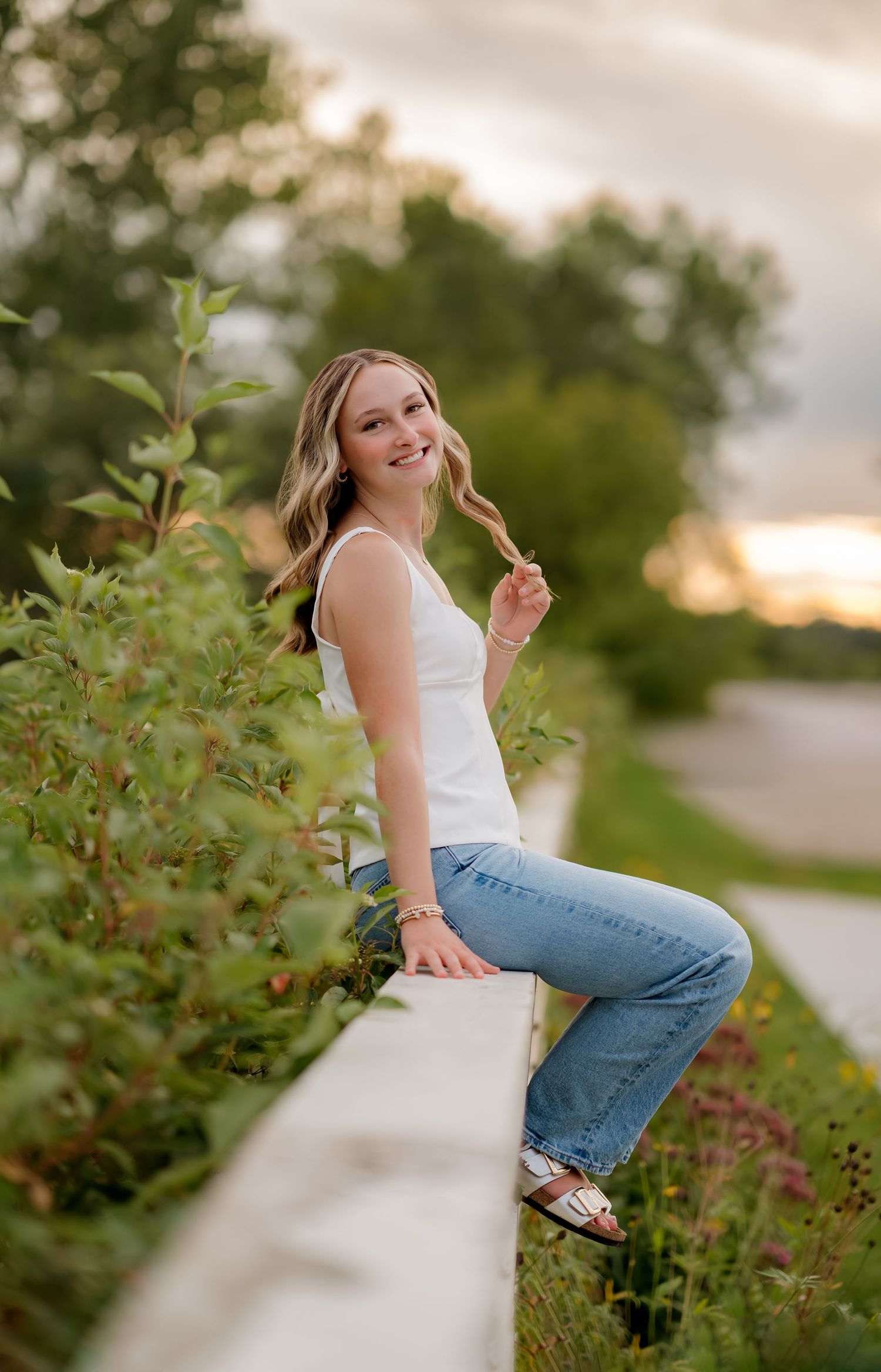 Young woman in white tank top and jeans sits on a white fence surrounded by lush greenery at golden hour.
