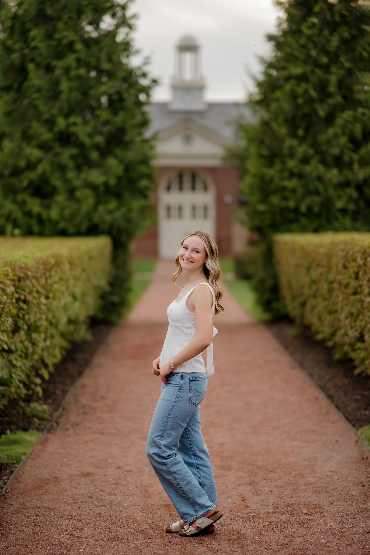 Smiling blonde woman in white tank and jeans poses on garden path with brick building behind.